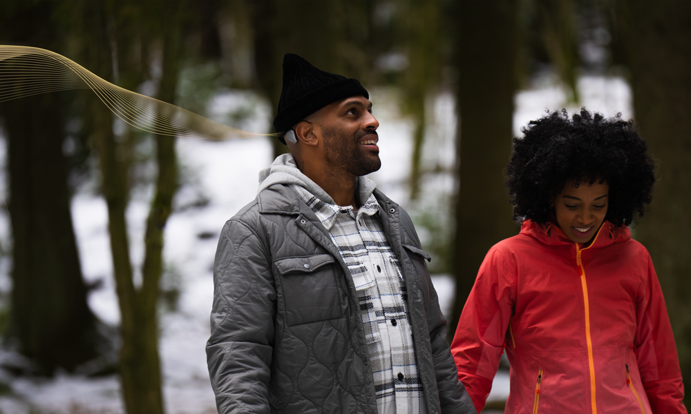 A couple laughing while taking a walk through a forest, with the man wearing a hat and a Sentio 1 Mini sound processor.