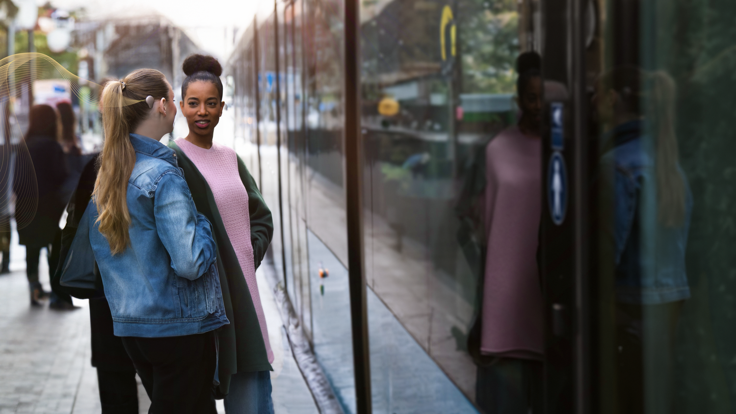 Two friends engaging in conversation at a bus stop, with one of them wearing a bone anchored hearing device.